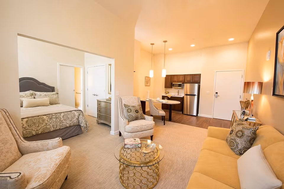 A bright and cozy senior living apartment featuring a living room with a yellow sofa, patterned armchairs, a glass coffee table, and a small dining area with two chairs. The kitchen area has wooden cabinets, a stainless steel refrigerator, and a microwave. The bedroom is partially visible with a bed dressed in patterned bedding and a gray upholstered headboard. The walls are painted in warm tones, and the space is well-lit with ceiling lights and pendant lamps.