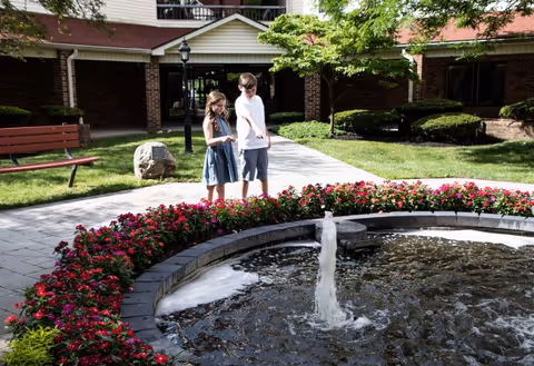 Two children, a girl and a boy, stand near a circular water fountain surrounded by red flowers in an outdoor garden area of a senior living facility. The background shows a brick building with windows, a bench, a lamp post, and green trees and shrubs.