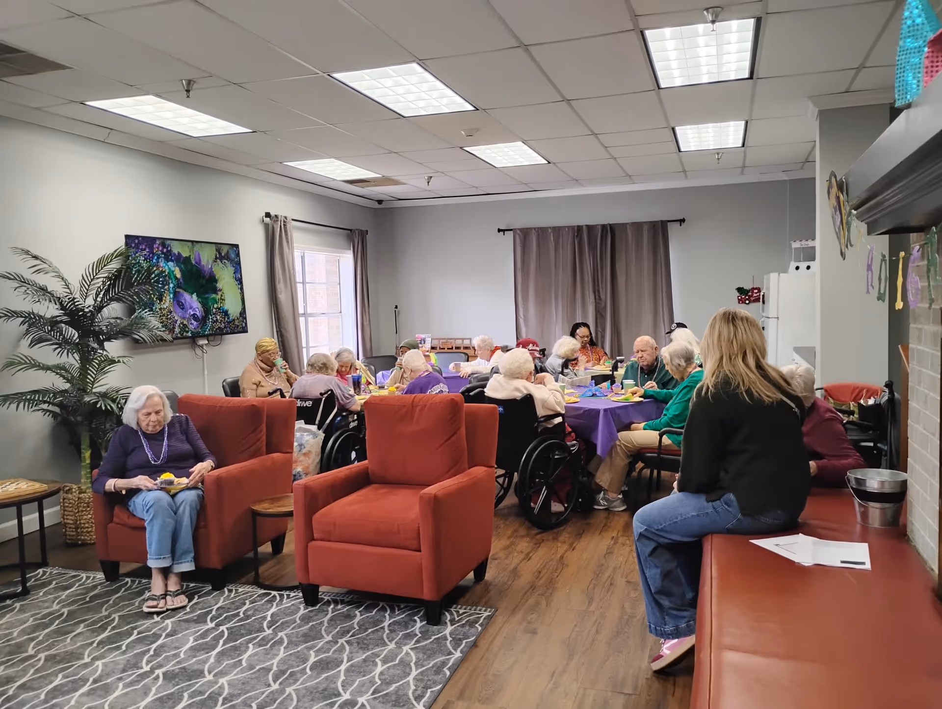 A group of elderly people sitting around a table covered with a purple tablecloth, eating and socializing in a common room. There are two red armchairs in the foreground, a woman sitting on one of them eating. A woman with blonde hair is sitting on a red bench on the right side. The room has a TV mounted on the wall, a large plant, and windows with curtains.