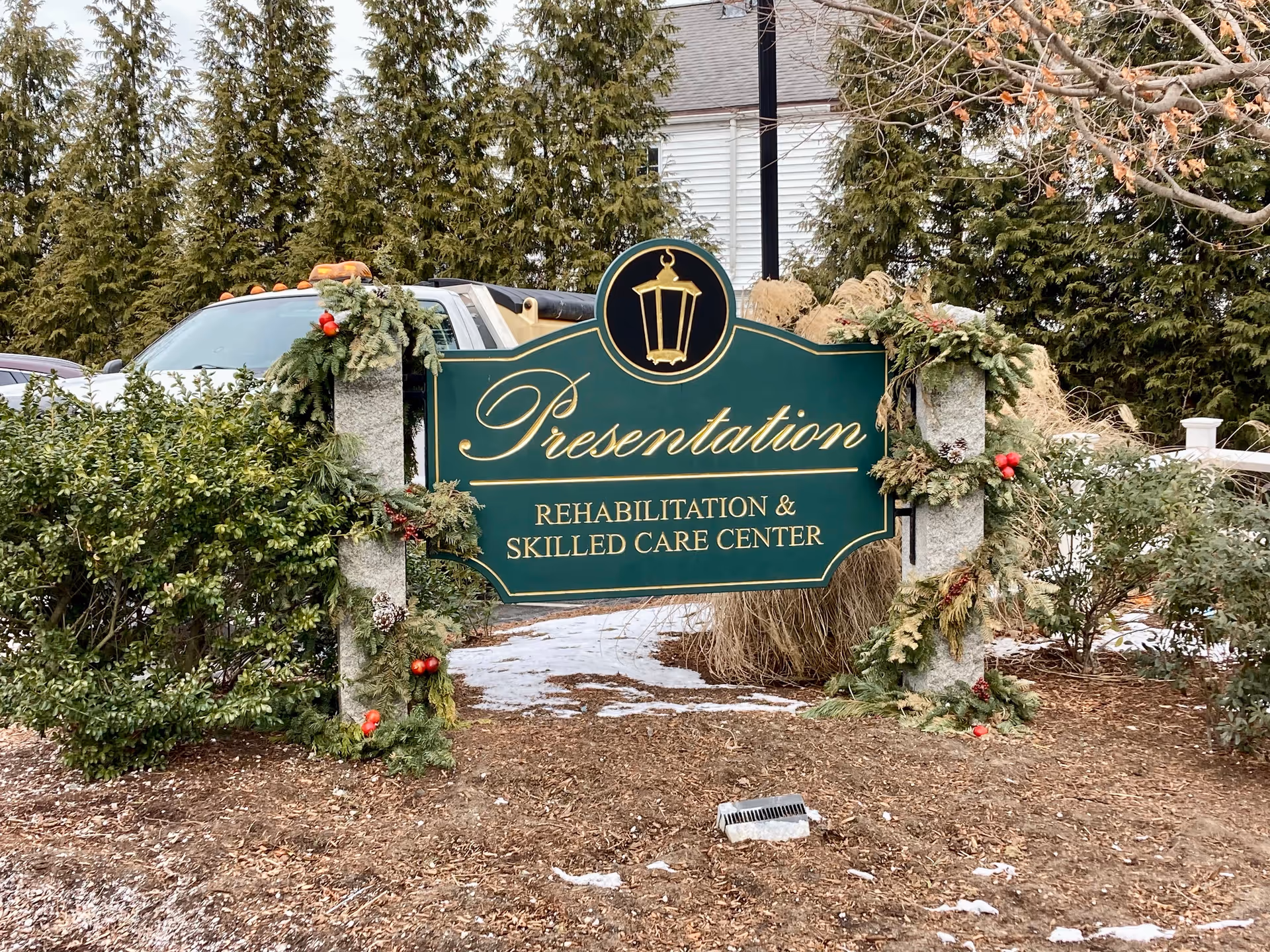 Outdoor sign for Presentation Rehabilitation & Skilled Care Center decorated with green garlands and red berries, surrounded by bushes and trees with some snow on the ground.