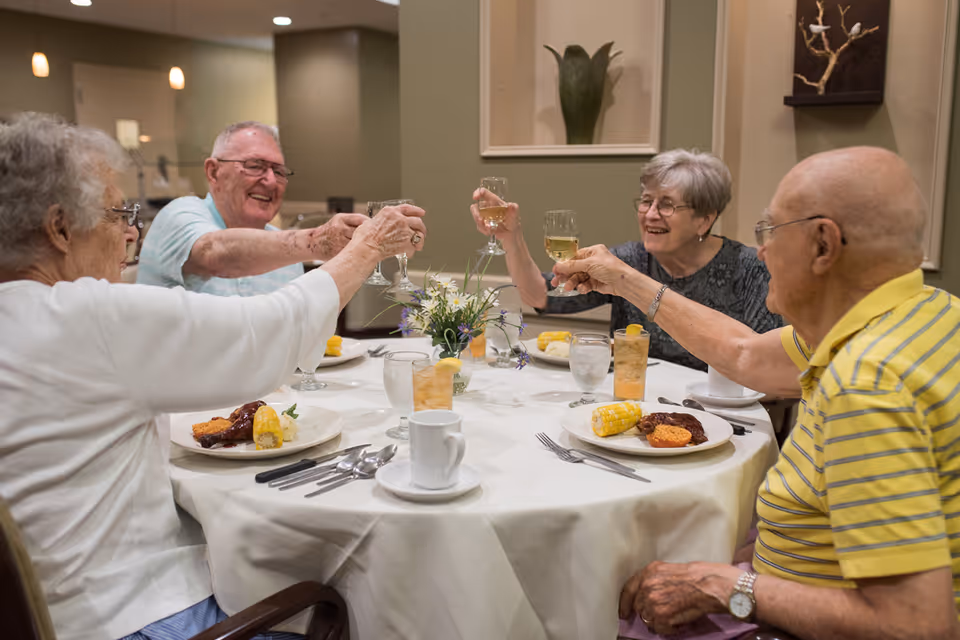 Four elderly people sitting around a round dining table in a senior living facility, raising their glasses in a toast. The table is set with plates of food including corn on the cob and a meat dish, glasses of iced tea, water, and coffee cups. There is a small floral centerpiece on the table and the room has a warm, inviting atmosphere.