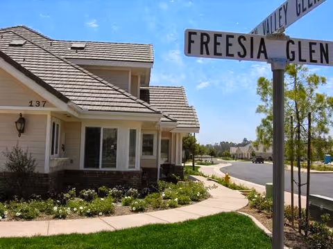 Front of a single-story retirement community home numbered 137 with landscaped lawns, a curved sidewalk and a street sign reading 'FREESIA GLEN' under a blue sky.
