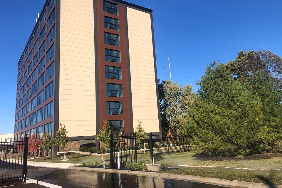 A modern senior living facility building with multiple floors, surrounded by greenery and a fence.