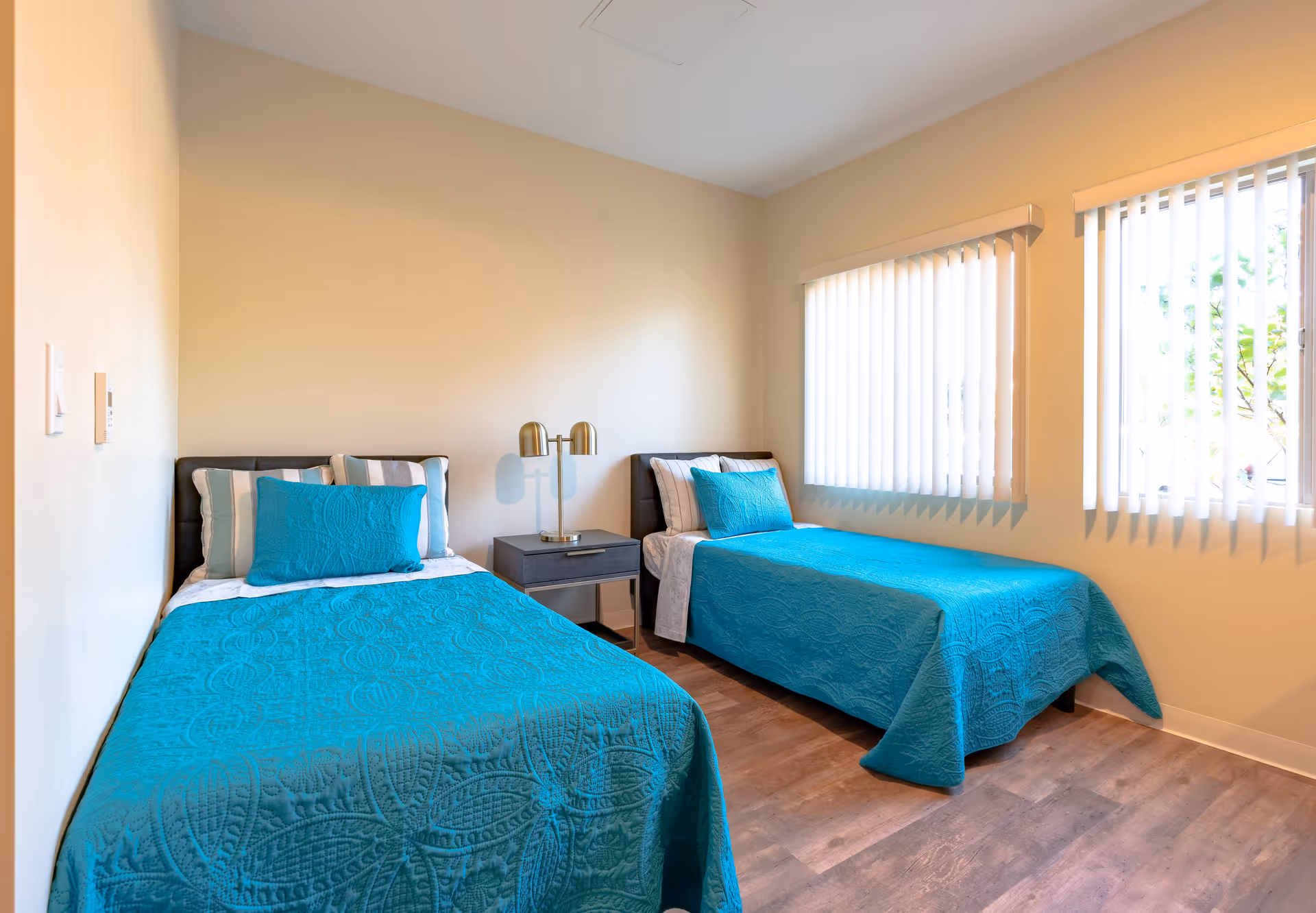 Two twin beds with teal bedspreads, a nightstand with a lamp, and windows with vertical blinds in a bright bedroom.