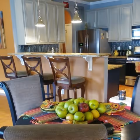 A dining table with a bowl of green apples in the foreground and a kitchen with bar stools, granite counter, stainless steel appliances, and white cabinets in the background.