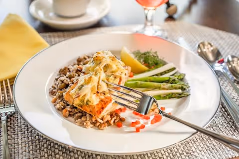 A plated meal consisting of a piece of baked fish topped with melted cheese and herbs, served on a bed of mixed grains with a side of asparagus, a lemon wedge, and garnished with diced red peppers. The plate is on a woven placemat with a fork resting on the plate, and a cup and a glass of pink beverage are visible in the background.