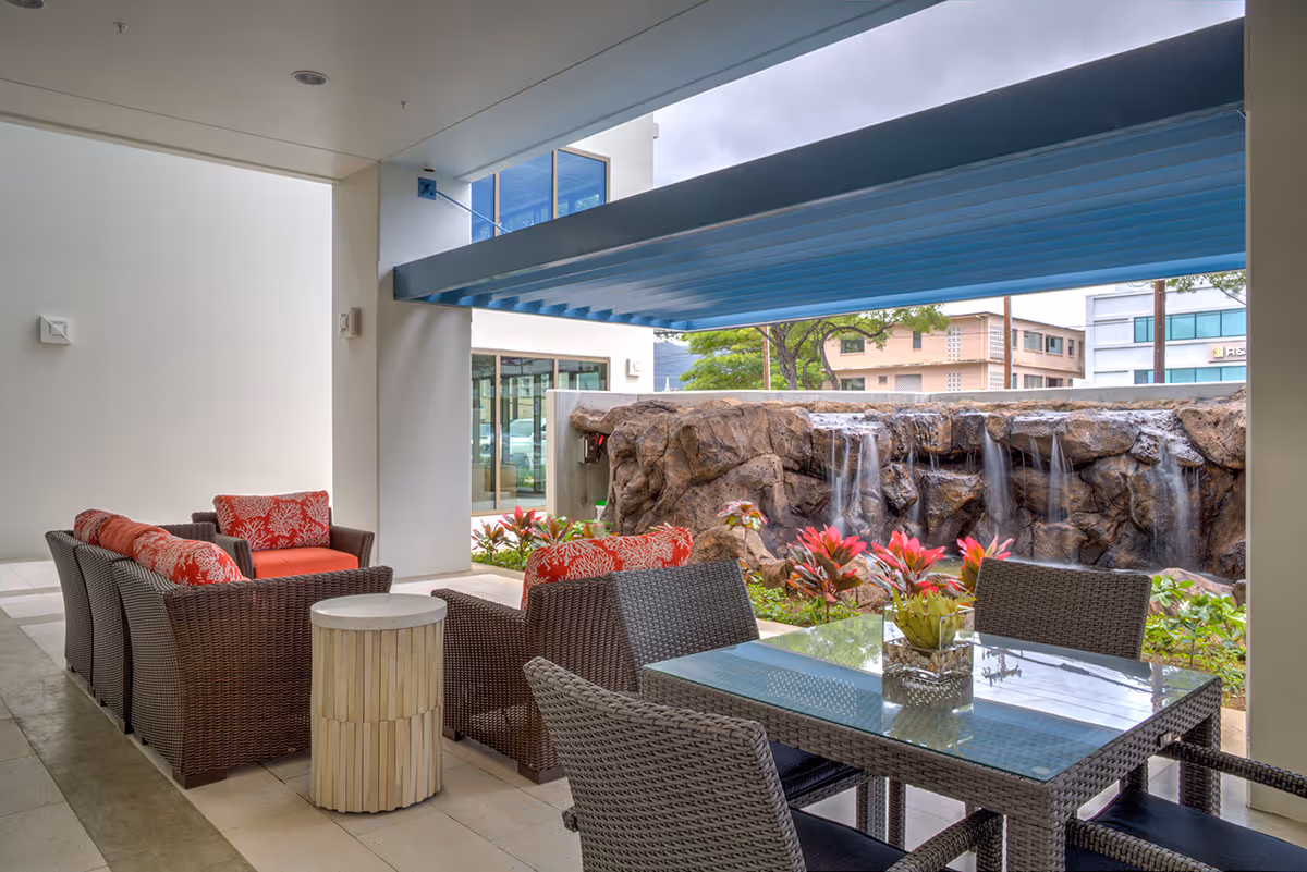Covered outdoor seating area with wicker chairs and red cushions, a glass-top table with chairs, and a small round side table. In the background, there is a rock wall with multiple small waterfalls and some red and green plants. The area is adjacent to a building with large windows.