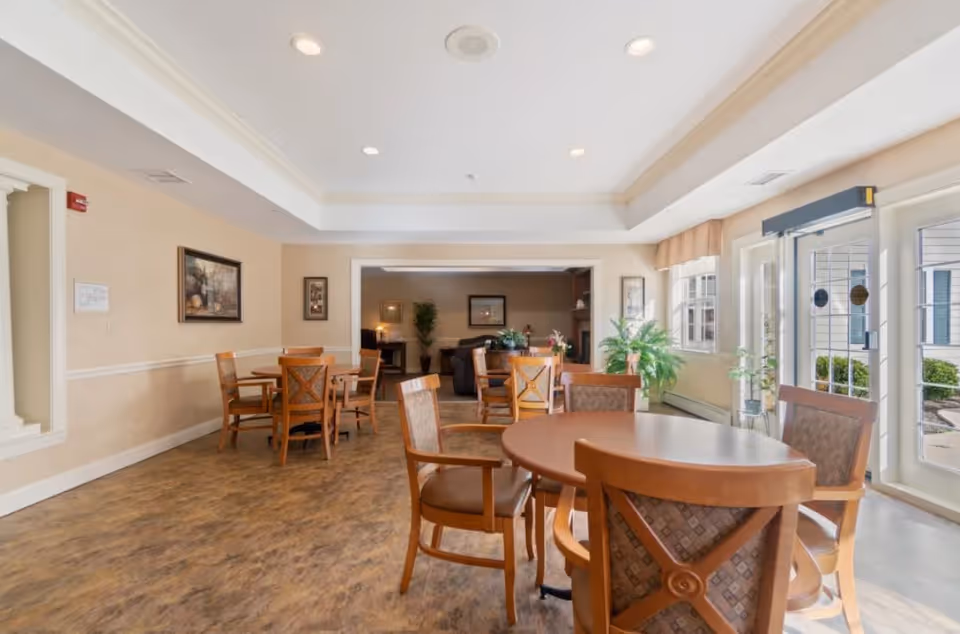 A bright and spacious common area in a senior living facility with several round wooden tables and chairs arranged for seating. Large windows and glass doors on the right side let in natural light, and there are plants placed near the windows. The room has a beige color scheme with framed artwork on the walls and a view into an adjoining lounge area with sofas and more plants.