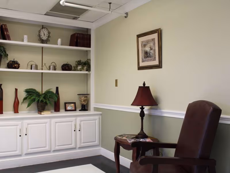 A cozy corner of a room with a brown armchair next to a small wooden side table holding a lamp with a red shade and some magazines. Behind the chair is a light green wall with a framed picture hanging. To the left, there is a white built-in shelving unit with various decorative items including plants, vases, books, and small sculptures.