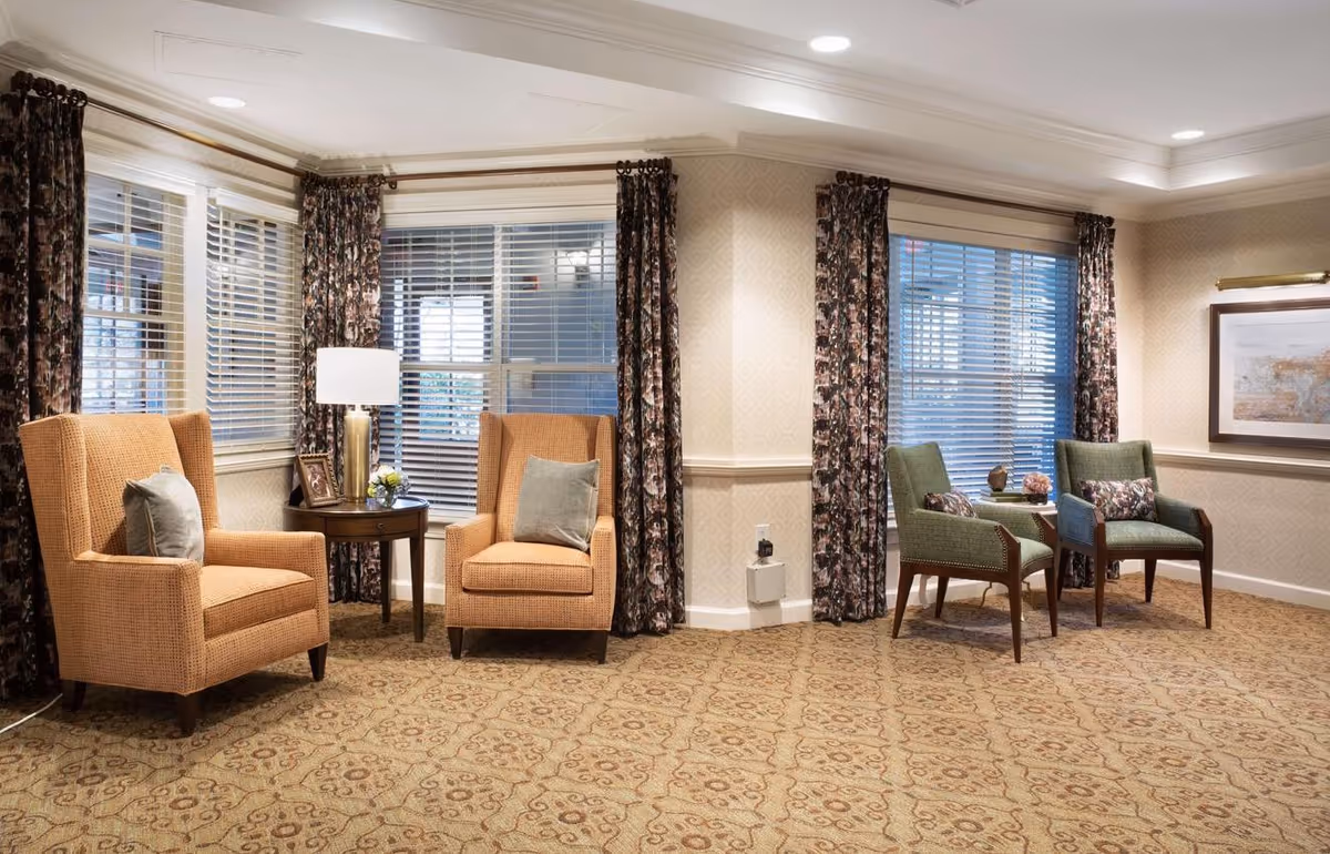 A cozy sitting area in a senior living facility with two orange armchairs and two green armchairs arranged near windows with floral curtains. A small round table with a lamp and decorative items is placed between the orange chairs, and another small table with decor is between the green chairs. The room has patterned carpet and soft lighting.