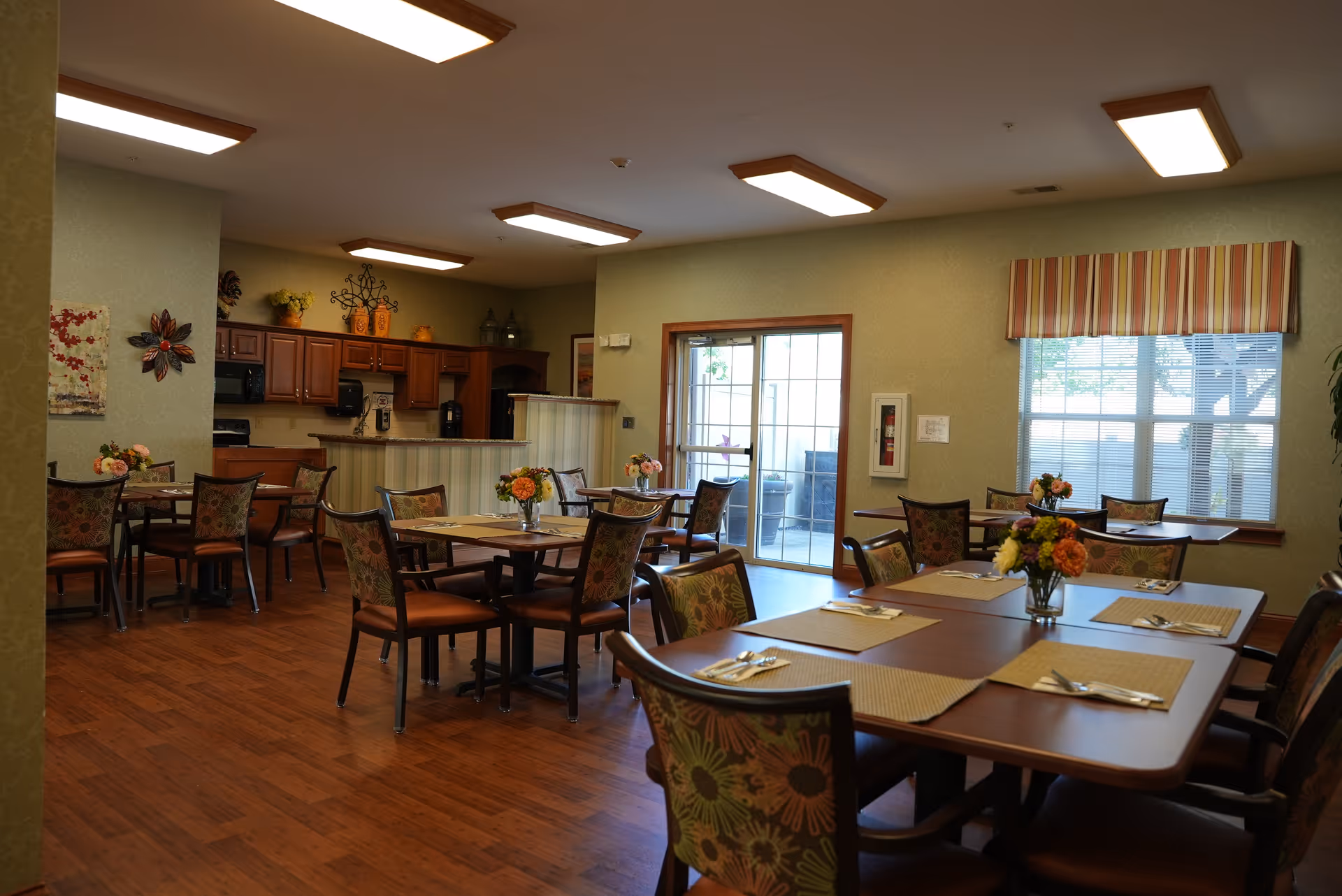 Bright dining room with multiple set tables and floral centerpieces, chairs, and a serving counter/kitchen area in the background.