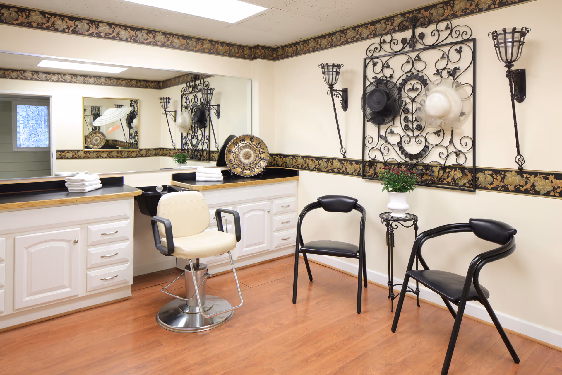 Interior view of a salon area with a beige salon chair in front of a counter with white cabinets and a large mirror. Two black chairs and a small table with a potted plant are positioned against the wall, which is decorated with a black wrought iron wall hanging holding two hats and two wall sconces. The floor is wooden and the walls have a floral border near the ceiling.
