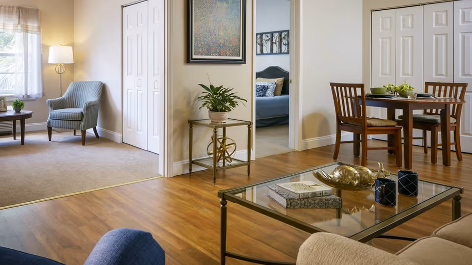 A cozy living space with wooden flooring, a glass coffee table with decorative items, a beige sofa, and a dining area with a wooden table and four chairs. To the left, there is a carpeted sitting area with a striped armchair, a side table with a plant, and a floor lamp near a window with sheer curtains. A doorway leads to a bedroom with a bed and framed artwork on the walls.