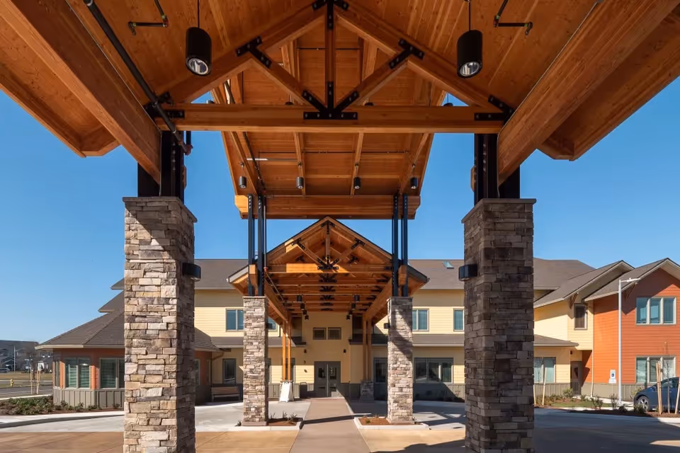 Entrance of Pear Valley Senior Living facility featuring a covered driveway with wooden beams and stone pillars under a clear blue sky.