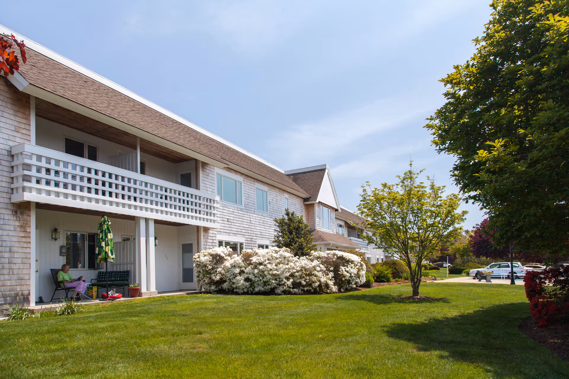 Exterior view of a two-story residential building with white siding and a brown roof. The building has balconies on the upper floor and patios on the ground floor. There is a well-maintained lawn with bushes and trees in front of the building. A person is sitting on a chair on one of the patios under a green umbrella. Several cars are parked in the background under a clear blue sky.