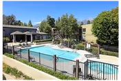 Outdoor swimming pool area at a senior living facility with lounge chairs, umbrellas, and a fenced perimeter. The pool is surrounded by trees and two-story buildings under a clear blue sky.