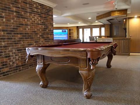 A spacious indoor recreational room featuring a wooden pool table with a red felt surface. The room has a brick accent wall on the left, carpeted floor, and a built-in wooden bar area in the background. Large windows allow natural light to enter the room.