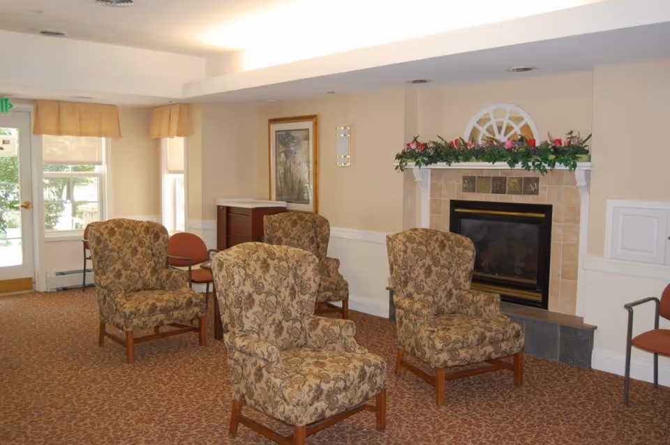 A cozy assisted living residence common area with four patterned armchairs arranged around a fireplace decorated with a floral garland. The room has beige walls, carpeted floor, a framed picture on the wall, and windows with beige valances letting in natural light.