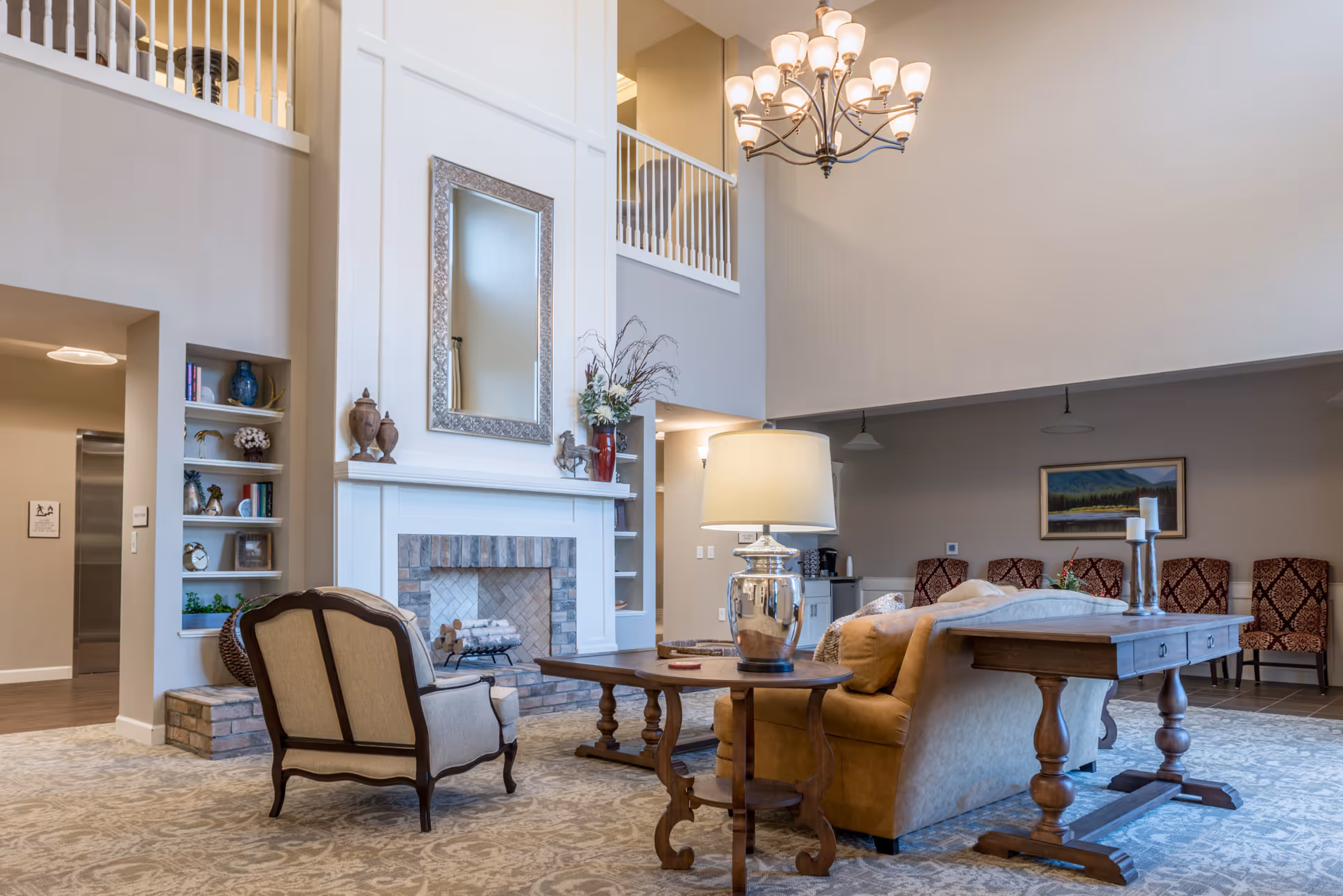 A spacious senior living facility common area with high ceilings, a large chandelier, and a fireplace with a decorative mirror above it. The room features a beige armchair, a brown leather sofa, wooden tables with a lamp and candles, and a patterned carpet. There are built-in shelves with decorative items and a painting on the far wall above a row of patterned chairs.
