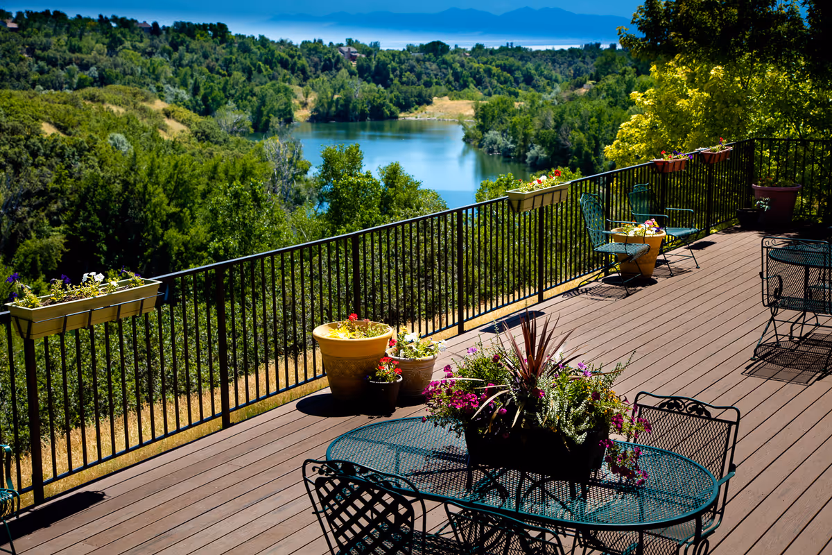 Wooden deck with metal patio tables and chairs, potted flowers, and railing overlooking a lake and tree-covered hills.