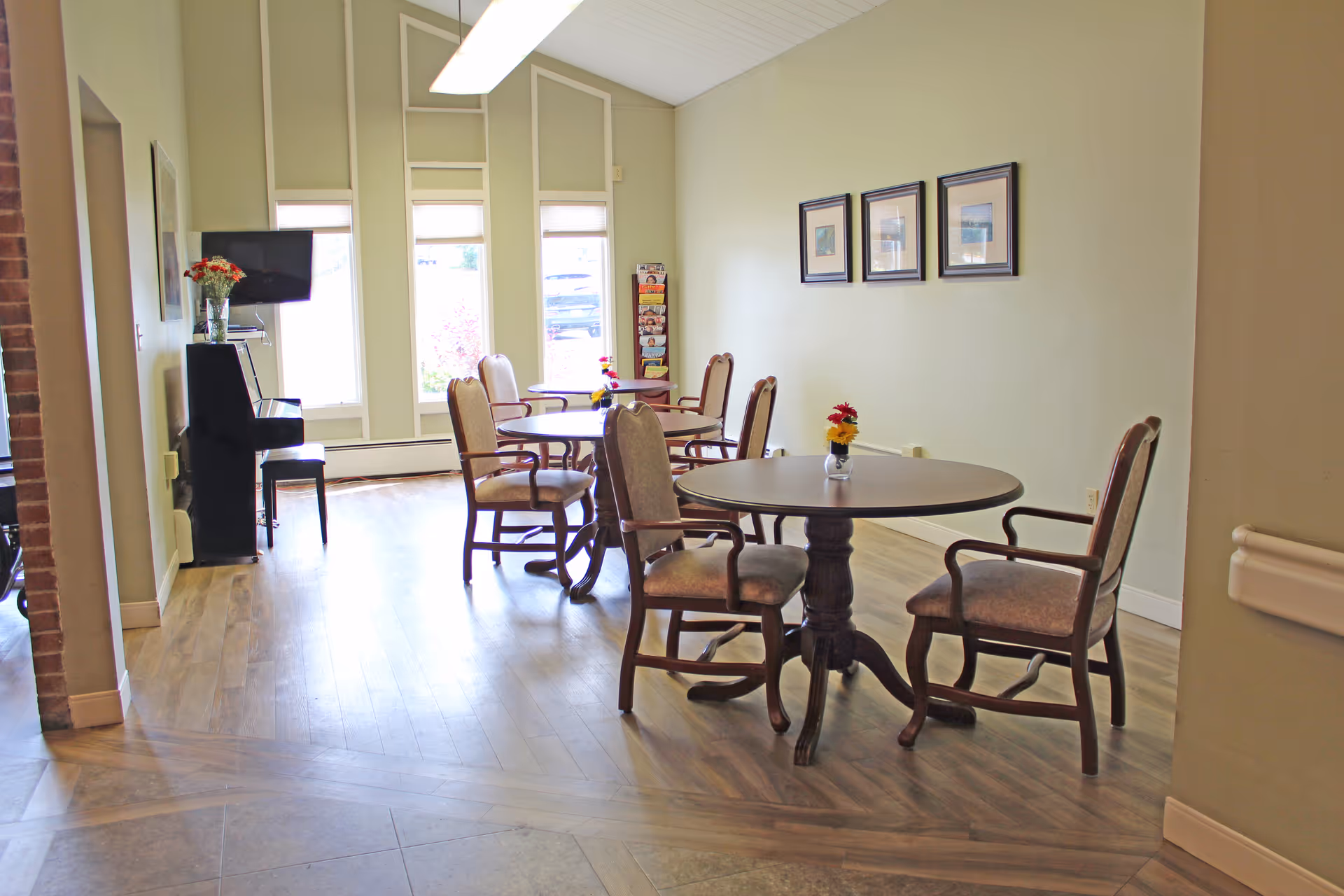 Bright communal dining/activity room with round tables, upholstered chairs, a TV and piano near tall windows.