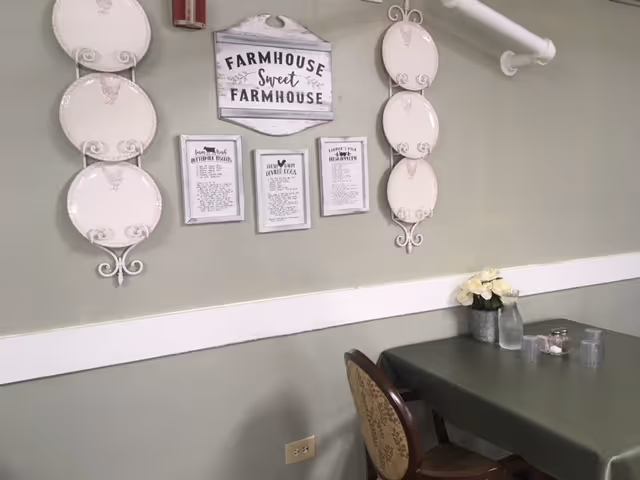 A dining area with a table covered by a green tablecloth, a chair with a patterned backrest, and a small flower arrangement in a metal vase. On the wall above the table are decorative plates and framed signs, including one that says 'Farmhouse Sweet Farmhouse'.