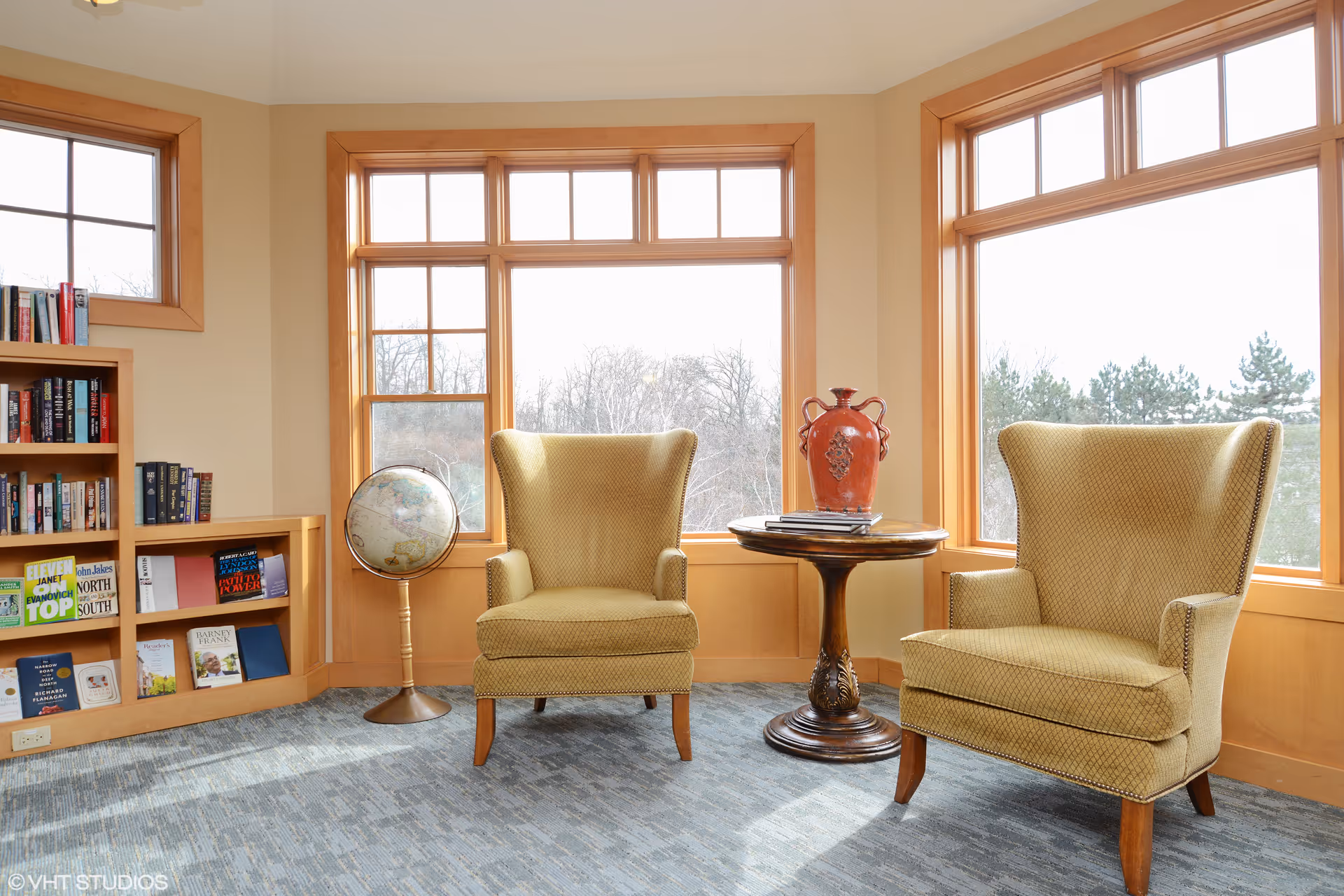 Sunlit seating area with two upholstered armchairs, a small round table with a vase, a globe, and a bookshelf by large windows.