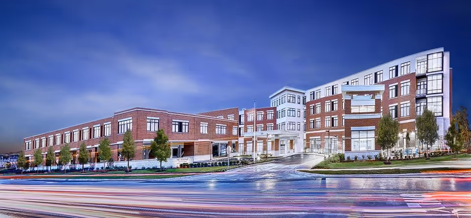 Exterior view of a large, modern multi-story senior living facility building with brick and white paneling, surrounded by landscaped greenery and trees under a blue sky at dusk.