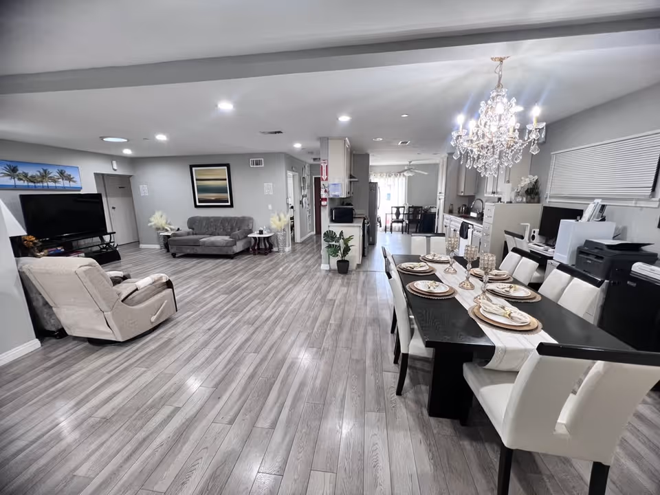 Open bright communal living and dining area with a chandelier-lit dining table, lounge seating, TV, and kitchen in the background.