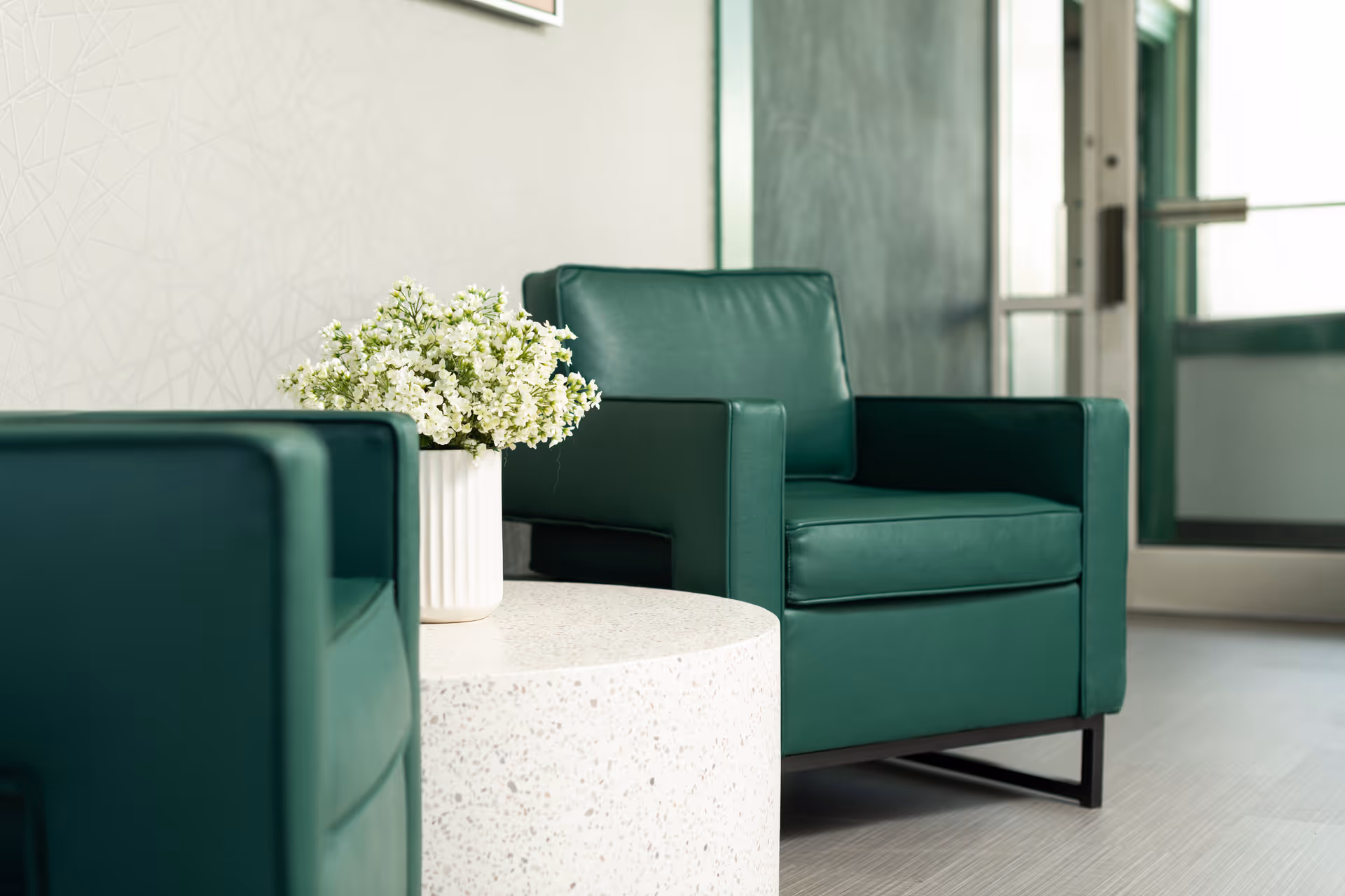 Two green armchairs positioned around a white round table with a white vase holding a bouquet of small white flowers, set in a bright room with a glass door in the background.