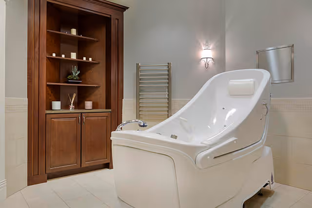 A modern, white therapeutic bathtub with a built-in headrest in a clean bathroom setting. To the left, there is a wooden cabinet with shelves holding candles and decorative items. A wall-mounted towel warmer and a light fixture are visible on the wall behind the tub.