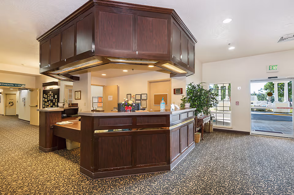 Reception area inside Inland Point Retirement Community featuring a dark wood front desk with a countertop, a patterned carpet floor, potted plants, and a glass exit door leading outside. The area is well-lit with ceiling lights and has a hallway leading to apartments.