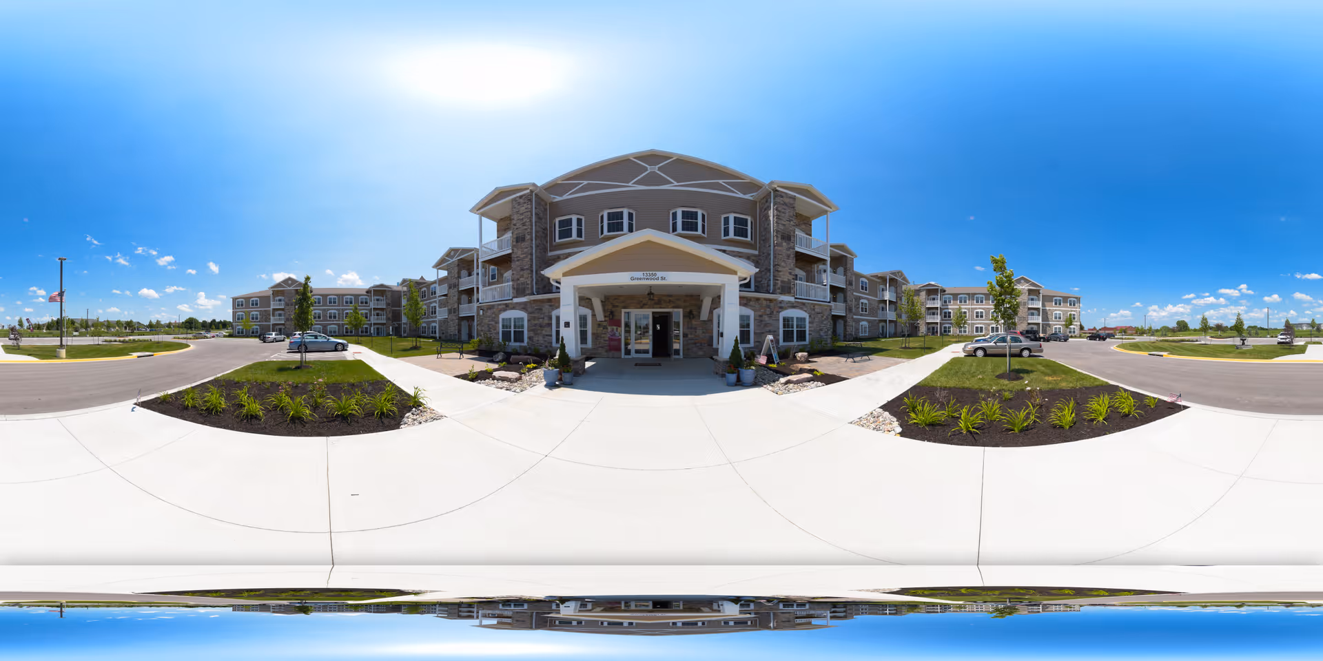 Front entrance of a large three-story senior living building with a covered porte-cochère, landscaped beds, and a circular driveway under a bright blue sky.