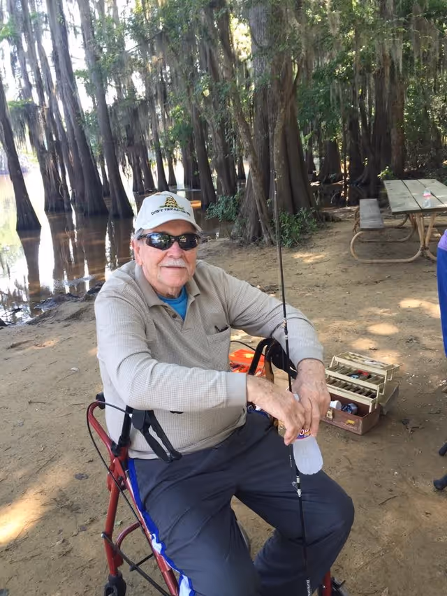An elderly man wearing sunglasses and a white cap sits on a red walker by a wooded lakeside area. He is holding a fishing rod and a water bottle, with a tackle box and picnic table visible in the background.