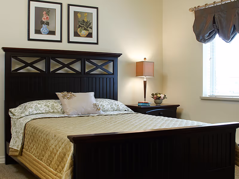 A neatly made bed with a dark wooden headboard and footboard in a bedroom. The bed has a beige quilted bedspread, floral pillowcases, and a decorative pillow. On the right side, there is a nightstand with a lamp, a small flower arrangement, and a couple of books. Two framed floral paintings hang on the wall above the headboard. A window with blinds and a brown valance is on the right wall.