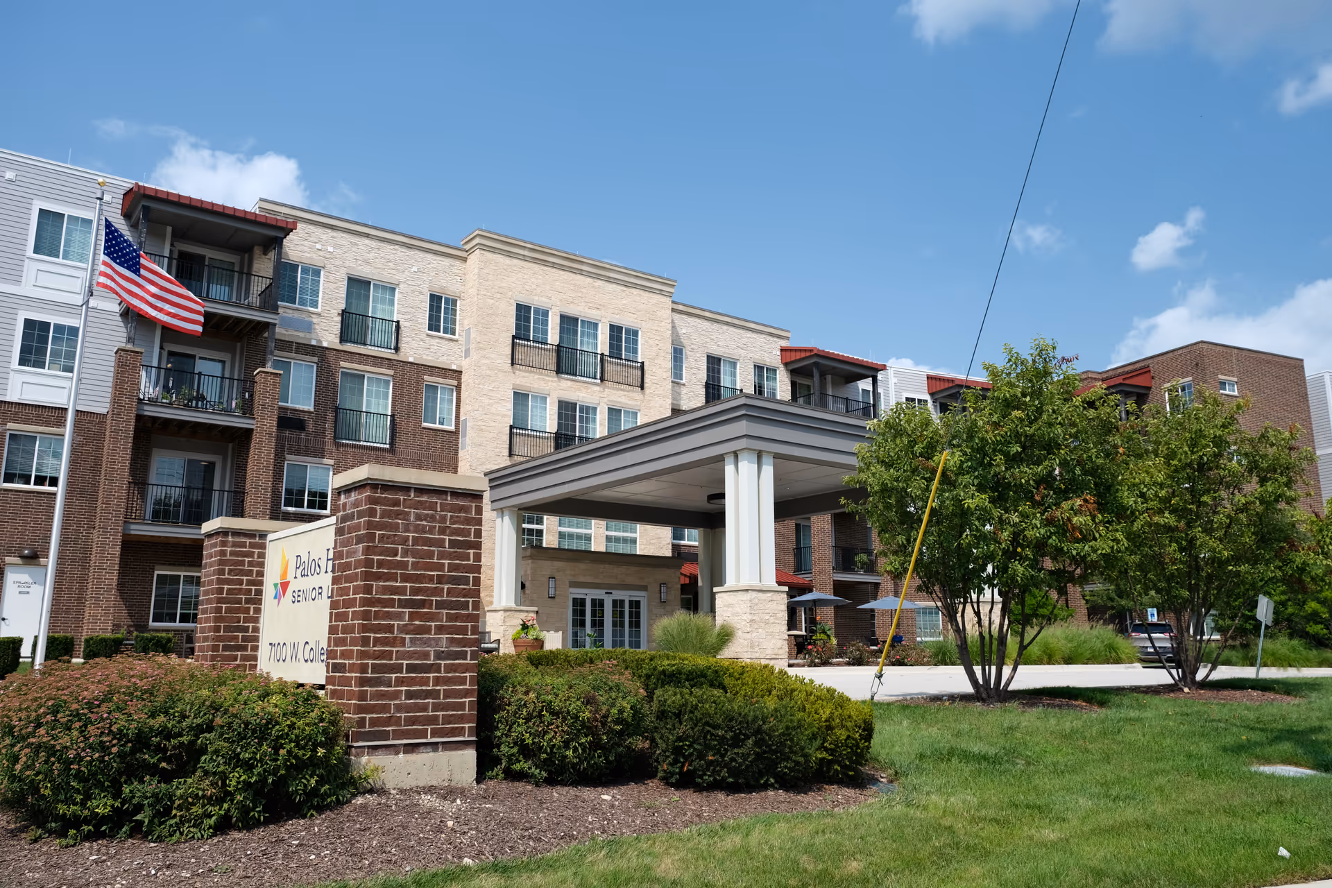 Exterior view of Palos Heights Senior Living facility showing a multi-story building with a covered entrance, brick and stone facade, balconies, an American flag, and landscaped greenery under a blue sky with some clouds.