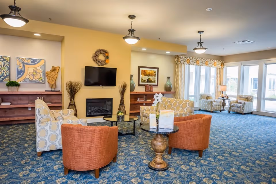 A spacious living room area with patterned blue carpet, multiple upholstered chairs and sofas in orange and beige tones, a wall-mounted flat screen TV above a fireplace, decorative vases, framed artwork, and large windows letting in natural light.