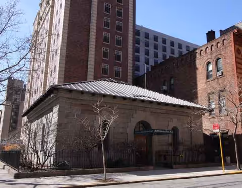 Front exterior of a stone building with a metal roof beside taller brick structures.