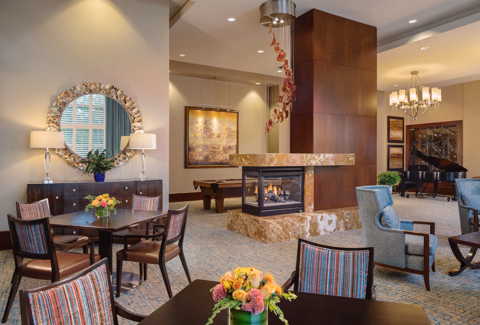 A cozy and elegant common area in a senior living facility featuring a central stone fireplace with a modern design. Surrounding the fireplace are several seating arrangements including wooden tables with striped cushioned chairs and comfortable armchairs. A decorative round mirror with a shell-like frame hangs above a dark wooden sideboard with two lamps and a plant. In the background, there is a pool table, framed artwork on the walls, a grand piano, and a chandelier, all contributing to a warm and inviting atmosphere.
