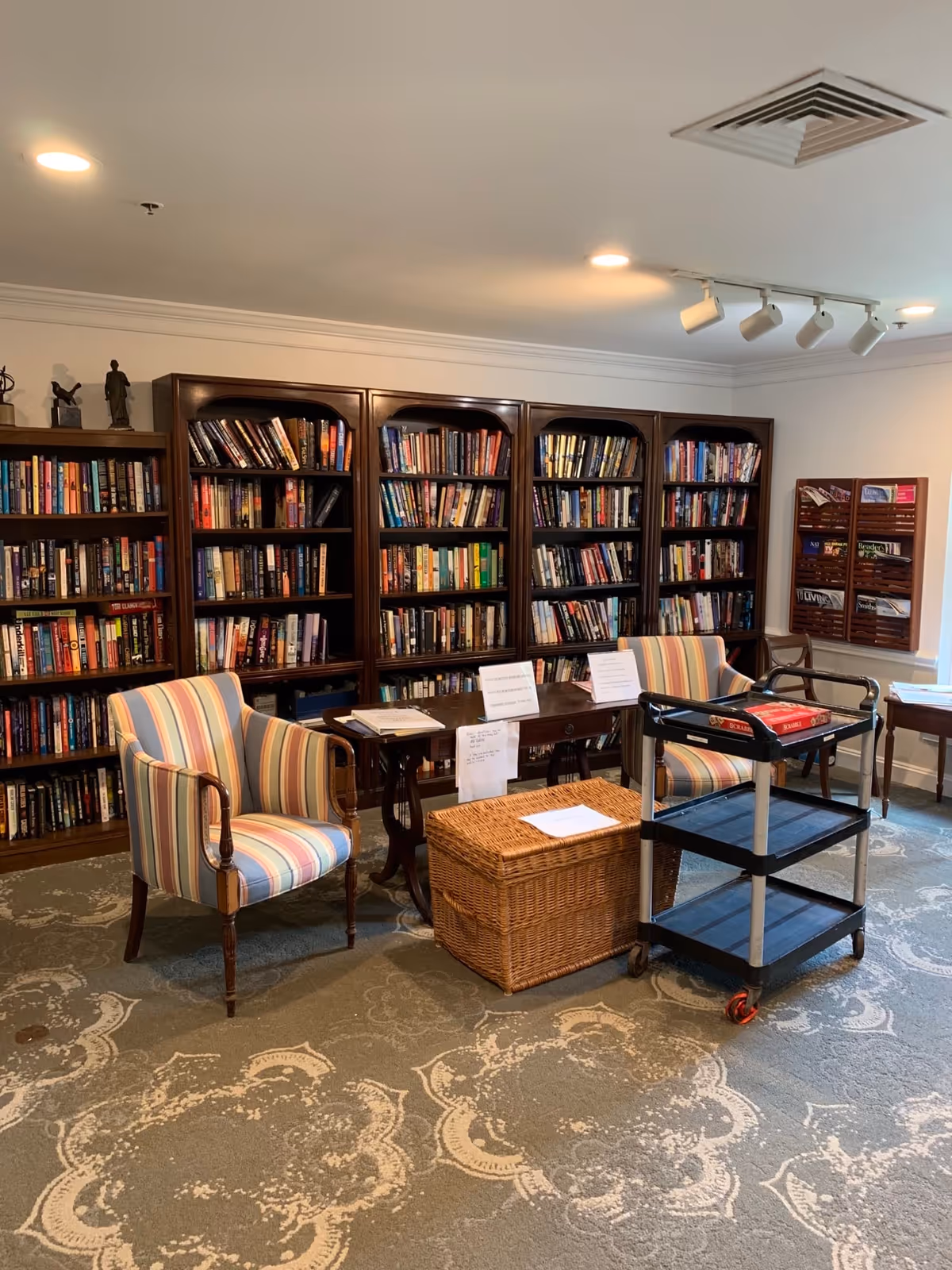 A cozy library room with several dark wooden bookshelves filled with books along the back wall. In front of the shelves are two striped upholstered armchairs, a wooden table with papers and signs on it, a wicker basket, and a black rolling cart with a Scrabble game on top. The room has a patterned carpet and white walls with ceiling lights.