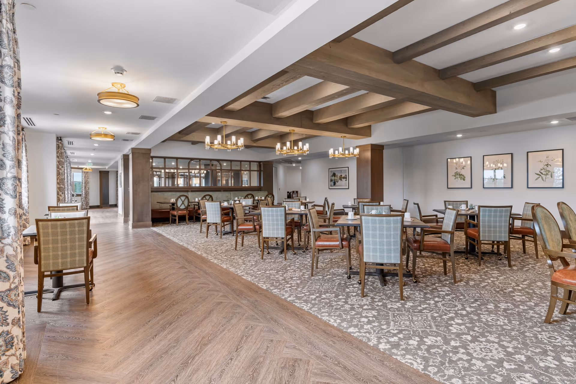A spacious dining room in a senior living facility with multiple tables and chairs arranged neatly. The room features a patterned carpet, wooden beams on the ceiling, chandeliers, and framed botanical artwork on the walls. Large windows with patterned curtains allow natural light to brighten the space.