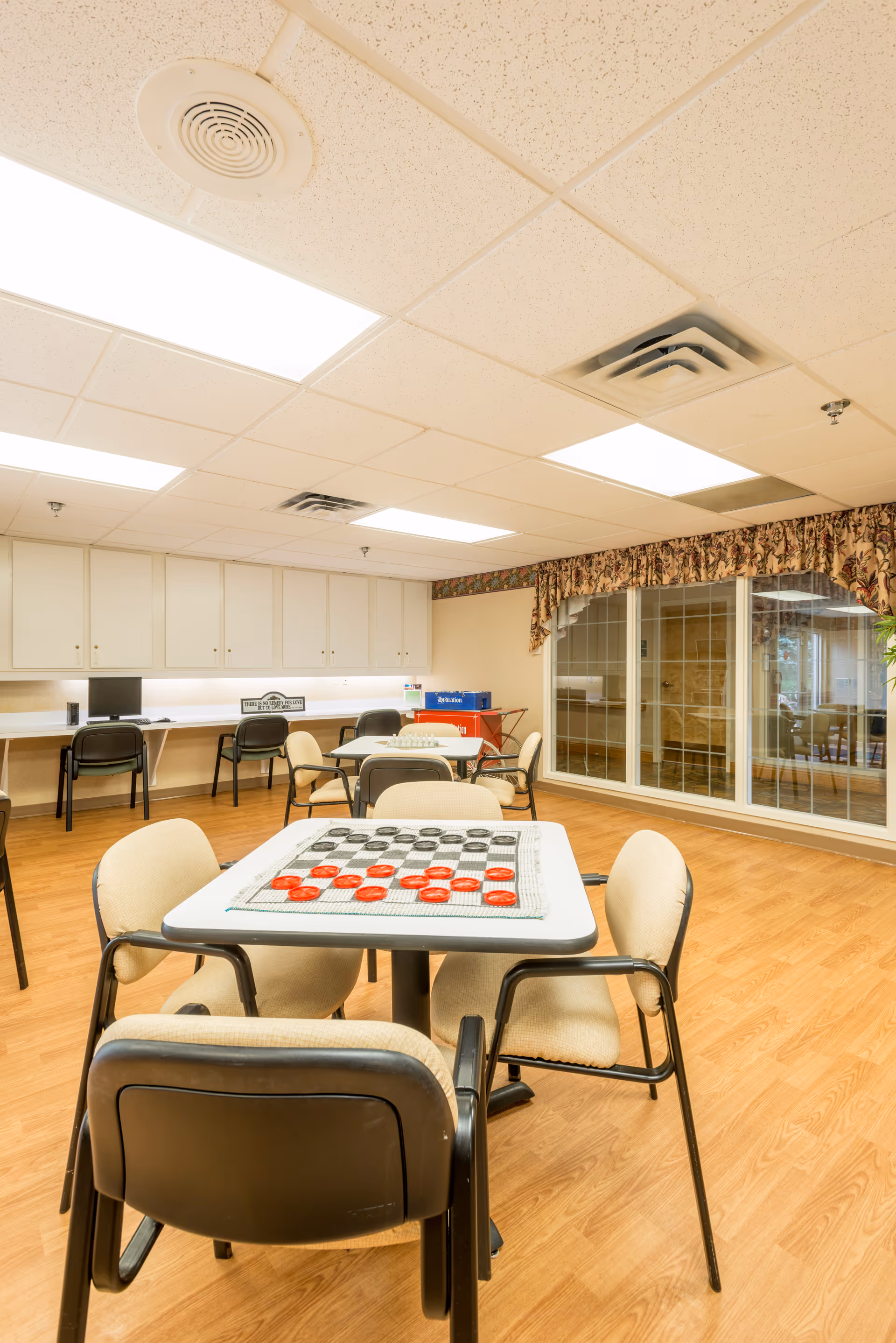 Activity room with tables and chairs, a checkers game on the front table, computer stations along the back wall and large windows.
