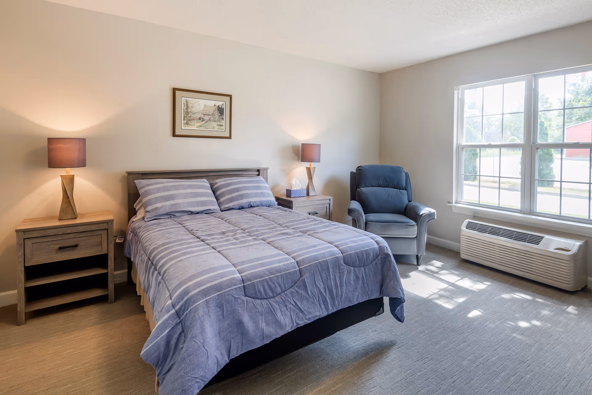 A tidy bedroom with a made bed, two bedside tables with lamps, an armchair by a large window, and an air conditioning unit.