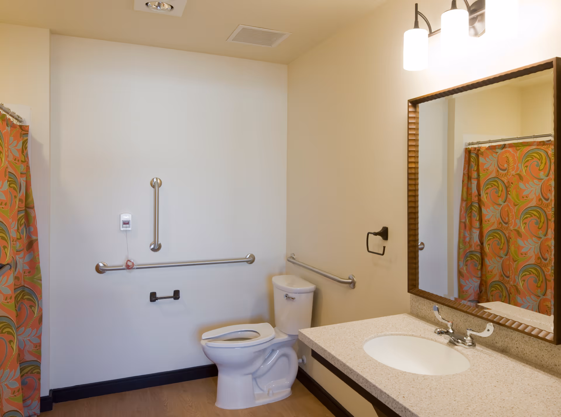 A clean bathroom with a toilet, a sink with a countertop, a large mirror above the sink, and a shower area with a colorful patterned shower curtain. The bathroom has grab bars installed near the toilet and on the wall, and a call button is visible on the wall next to the toilet.