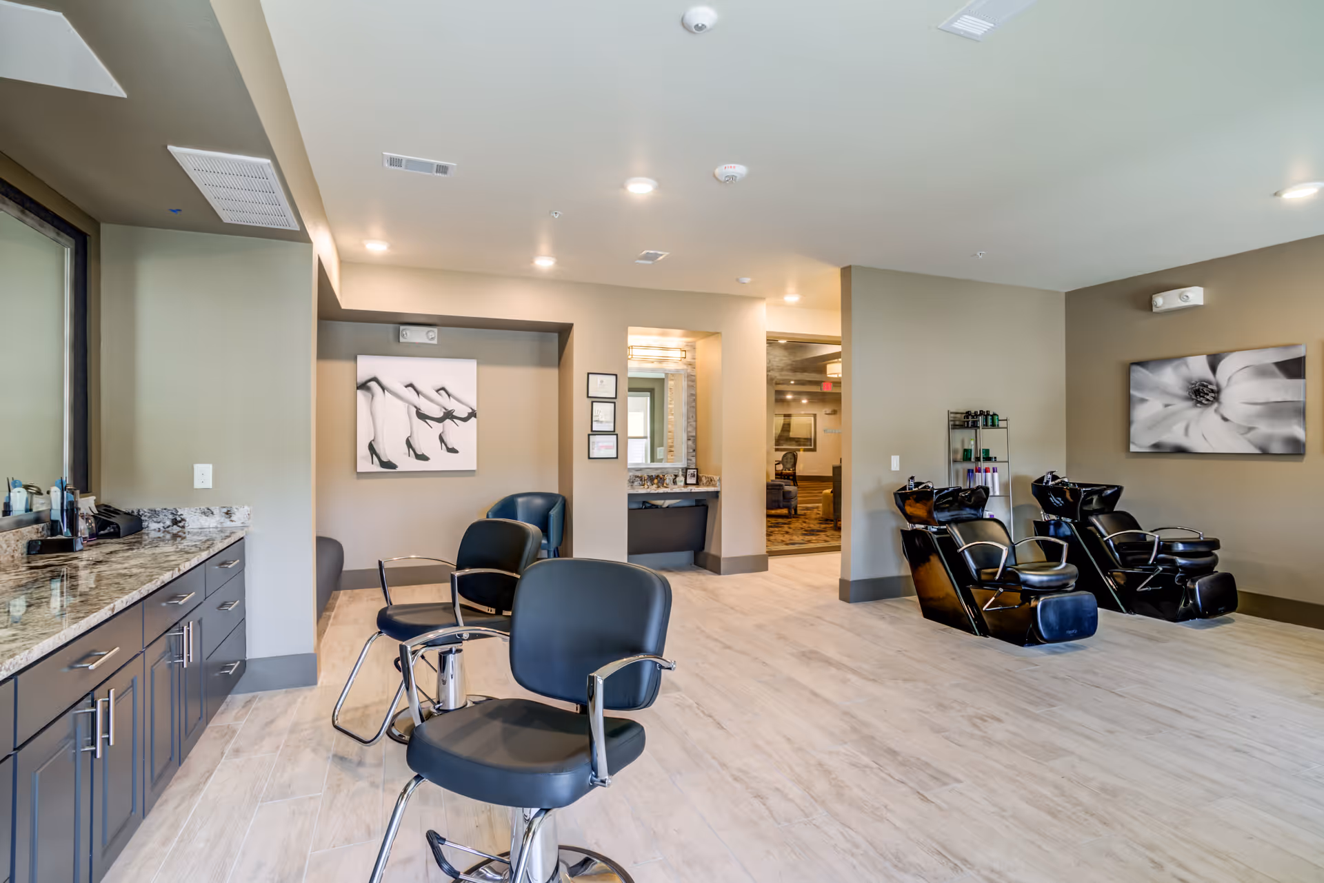 Interior view of a modern hair salon area in a senior living facility with styling chairs, hair washing stations, a large mirror, and decorative black and white artwork on the walls.