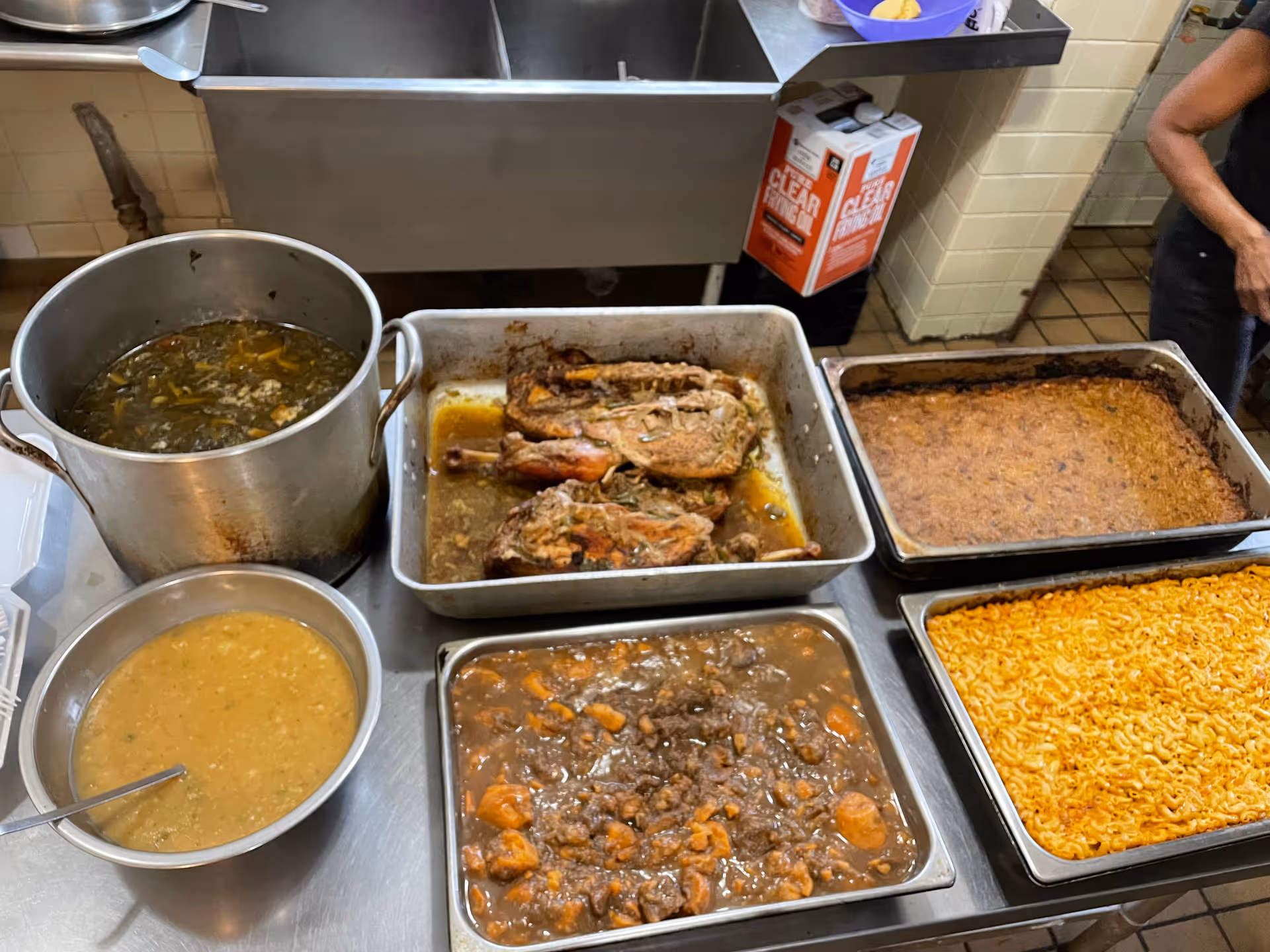 A kitchen counter with various cooked dishes including a large pot of soup or stew, a bowl of thick sauce or gravy with a spoon, a tray of roasted meat, a tray of baked casserole, a tray of macaroni and cheese, and a tray of beef stew with carrots. A person is partially visible on the right side, and kitchen equipment and supplies are in the background.