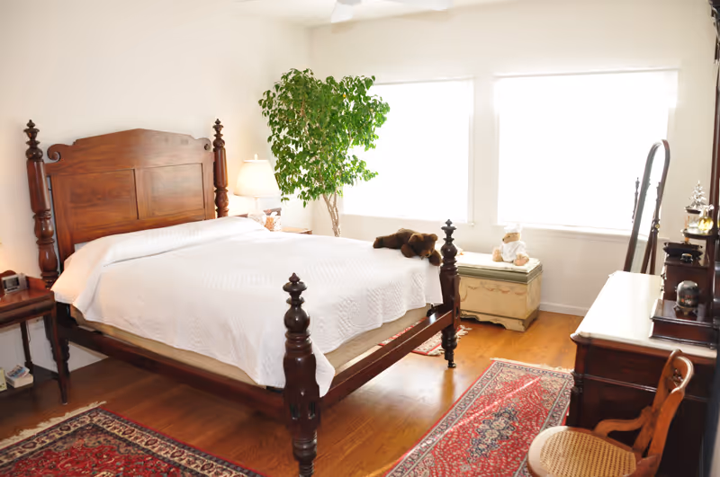 A bright bedroom with a wooden four-poster bed covered in a white quilt. There is a bedside table with a lamp and a clock, a large green potted plant near two large windows, a wooden chest with a stuffed animal on top, a wooden dresser with a mirror, and a wooden chair with a woven seat. The floor is hardwood with two red patterned rugs.
