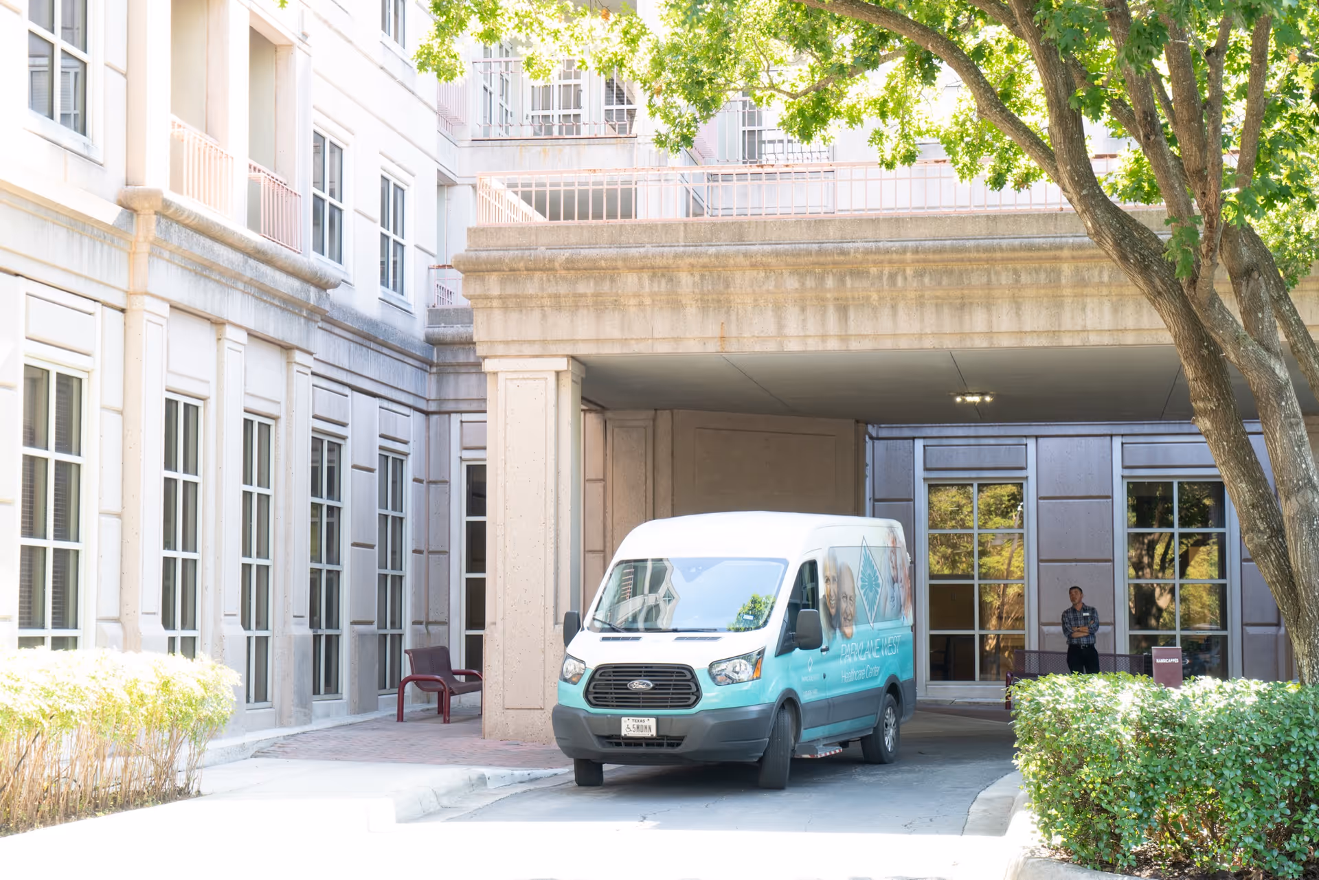 A white and teal van parked under a covered driveway at a multi-story building with large windows. A person stands near the building entrance, and there are trees and bushes around the driveway area.