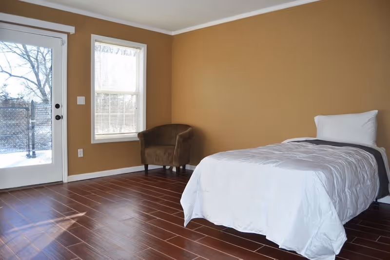 A simple bedroom with a single bed covered in white bedding, a brown armchair in the corner, a window with blinds, and a glass door leading outside. The walls are painted a warm tan color and the floor is dark wood.