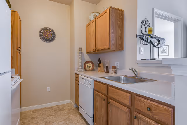 A compact kitchen with wooden cabinets, a white dishwasher, a stainless steel sink, and a white refrigerator. The countertop has a few decorative items including a candle holder and a small plate. A wall clock is mounted on the beige wall, and there is a pass-through window with a decorative metal shelf holding a candle.